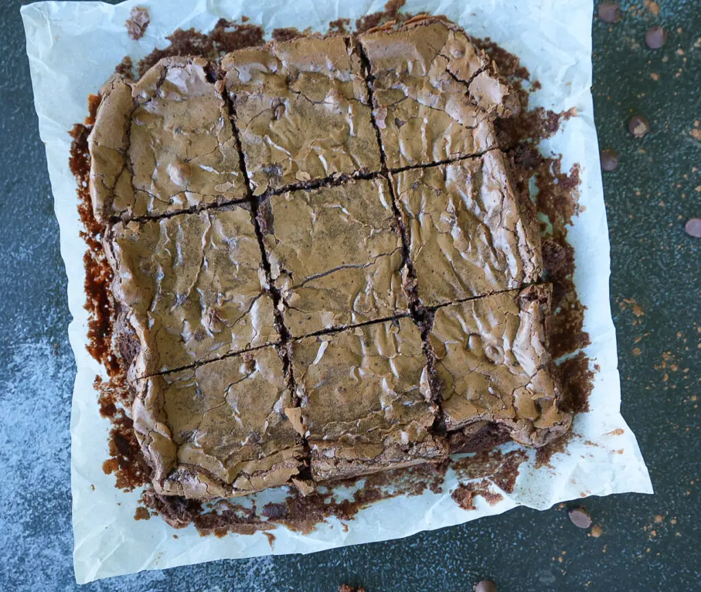 A tray of chocolate brownies cut into nine squares, resting on parchment paper with crumbs and a few chocolate chips scattered around.