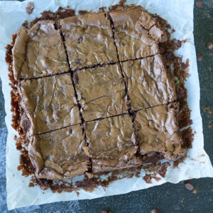 A tray of chocolate brownies cut into nine squares, resting on parchment paper with crumbs and a few chocolate chips scattered around.