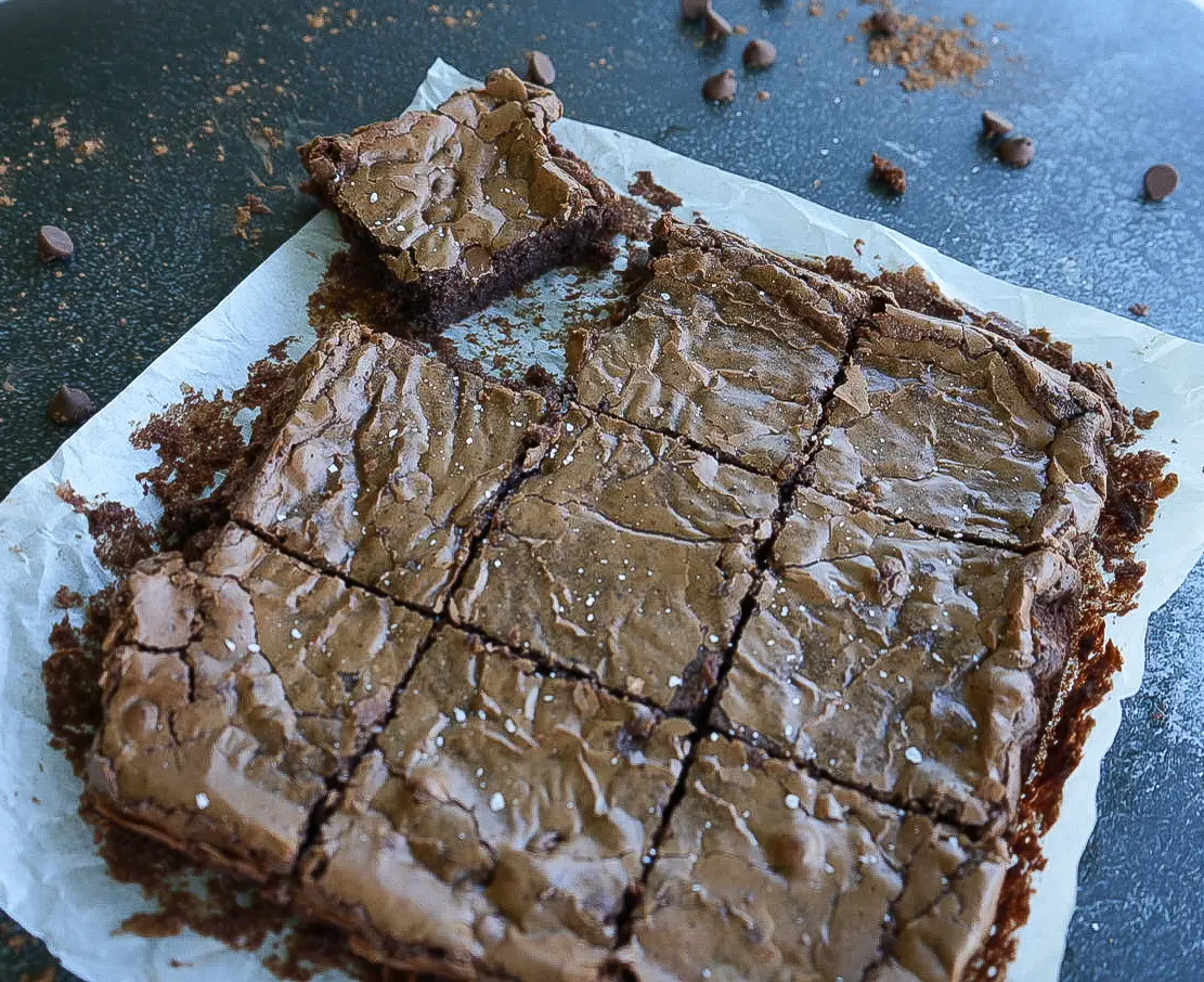 A batch of brownies cut into squares on parchment paper, with one brownie missing a bite. Chocolate chips are scattered around on the dark surface.