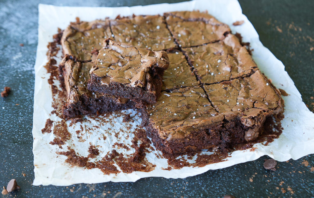 A batch of chocolate brownies on parchment paper, partly cut into squares with two brownies stacked in the center and crumbs scattered around.