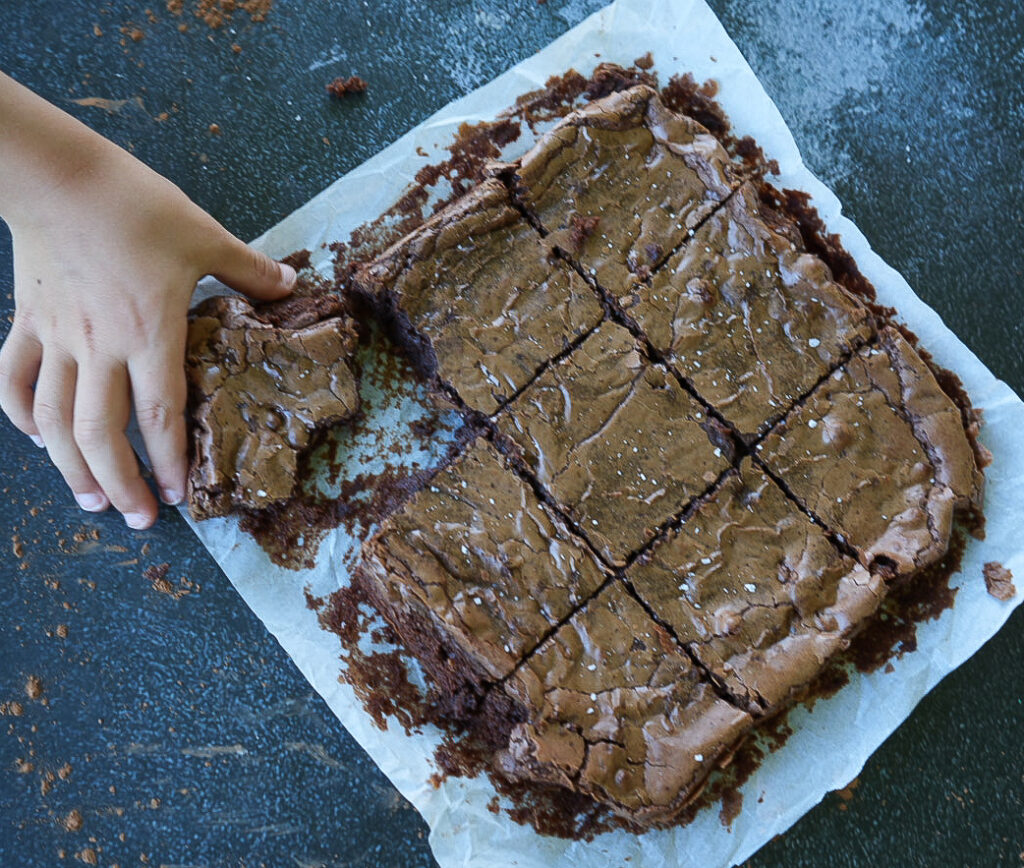 A hand grabbing a piece of chocolate brownie from a batch of brownies cut into squares on parchment paper.