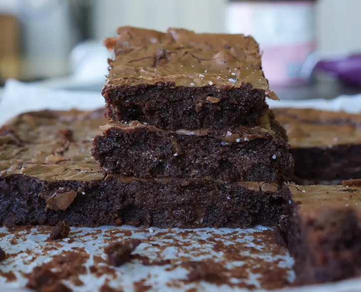 A stack of homemade sourdough discard brownies with a cracked top sits on parchment paper, surrounded by crumbs.