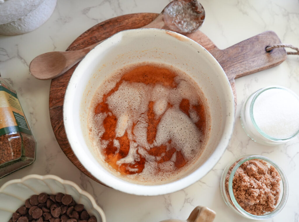 A bowl of melted butter with foam on top sits on a wooden cutting board, surrounded by sugar, brown sugar, chocolate chips, and a wooden spoon.