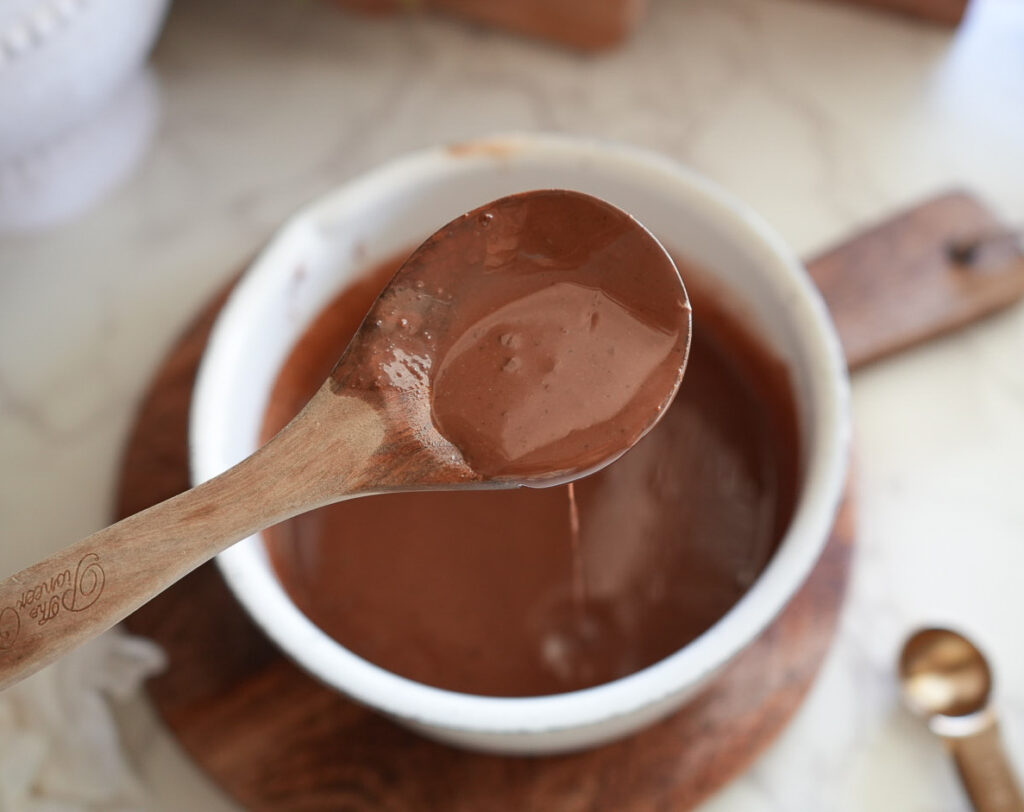 A wooden spoon holds thick chocolate sauce above a white bowl filled with more chocolate sauce, set on a wooden board.