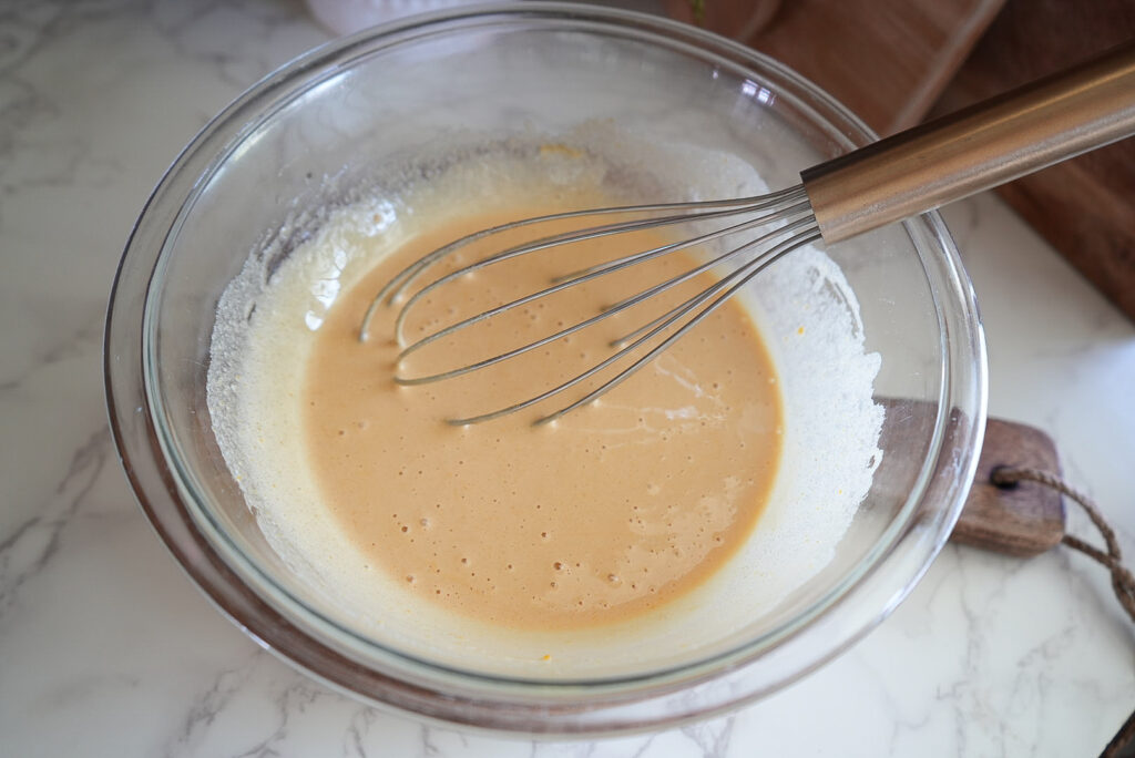 A glass bowl filled with a light brown batter and a metal whisk on a marble countertop.