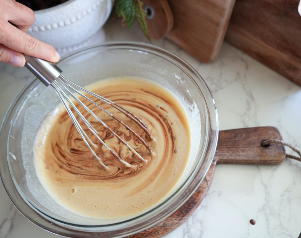 A hand uses a whisk to mix chocolate into a bowl of batter on a wooden cutting board on a marble countertop.