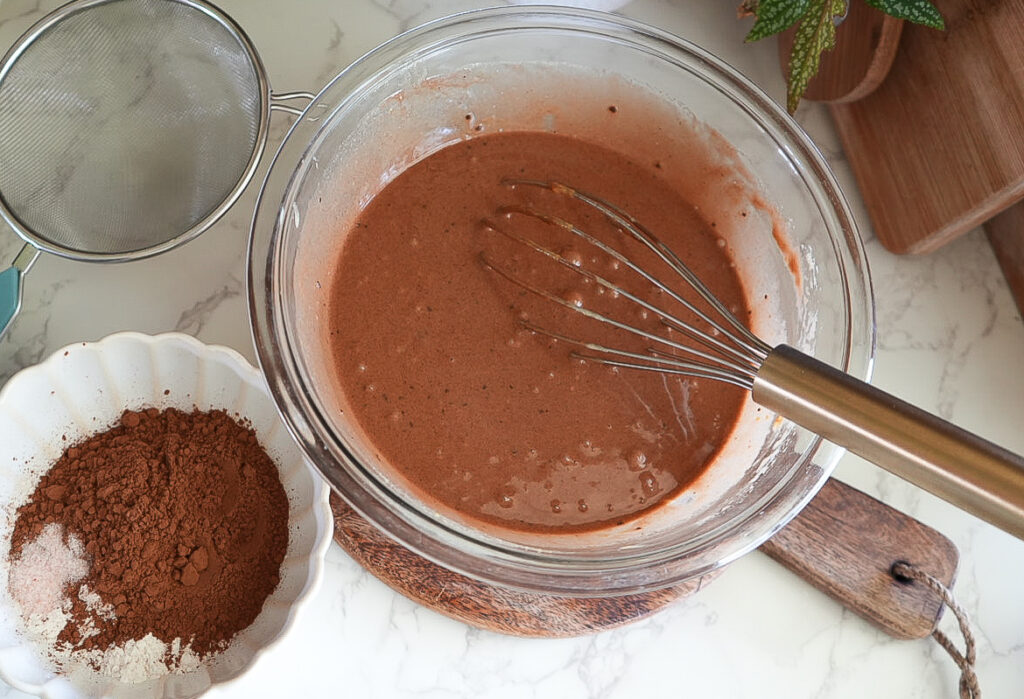 A glass bowl with chocolate batter and a whisk, next to a sieve and a dish containing cocoa powder and other dry ingredients on a kitchen counter.