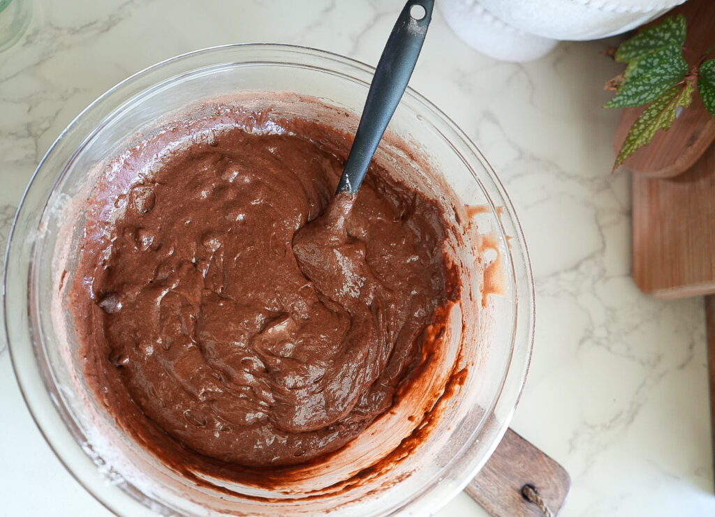 A glass bowl filled with chocolate cake batter and a black spoon on a marble countertop.