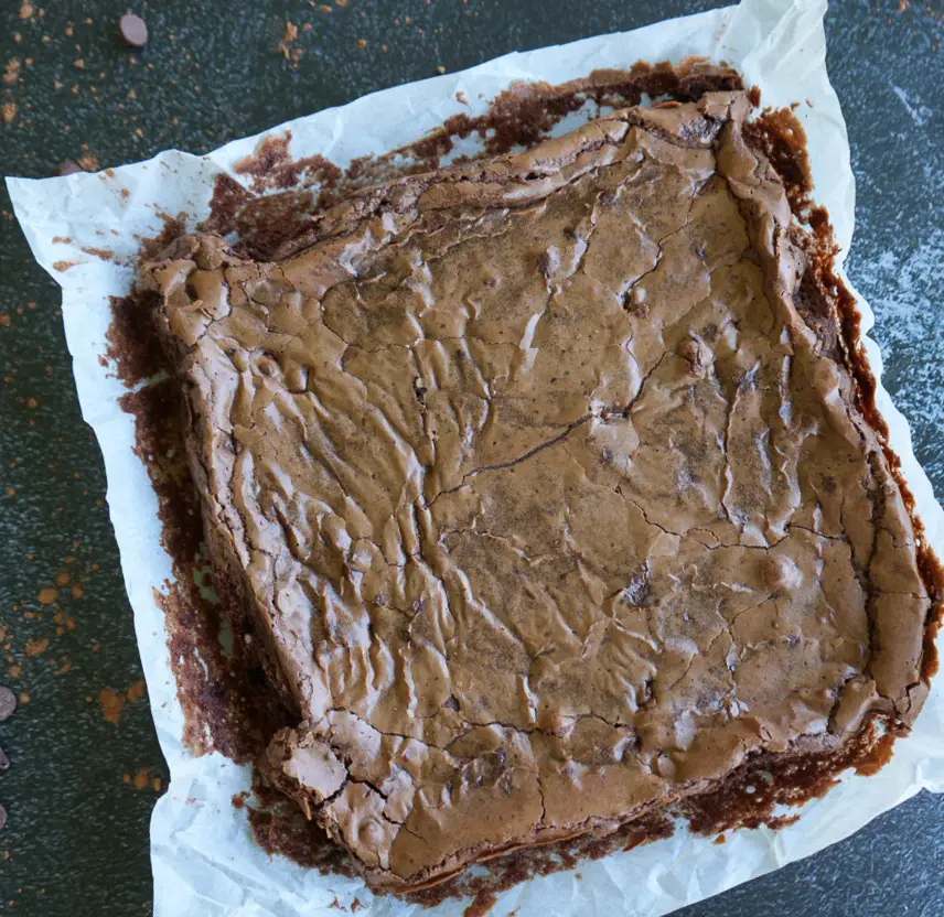 A baked tray of brownies on parchment paper, showing a cracked, flaky top and slightly uneven edges on a dark countertop.