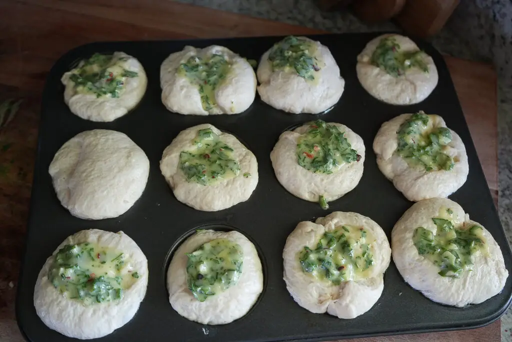 A muffin tin holds twelve rounds of dough, some topped with a mixture of chopped herbs and butter, ready to be baked.