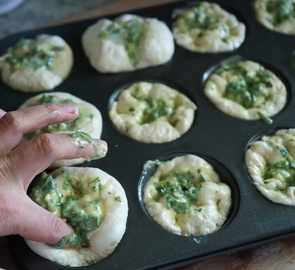 A hand presses dough topped with a green herb mixture into a muffin tin, preparing individual servings before baking.