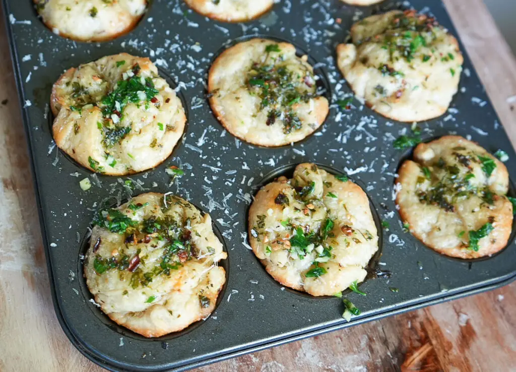 A muffin tin holds several baked savory biscuits topped with chopped herbs and grated cheese, resting on a wooden surface.