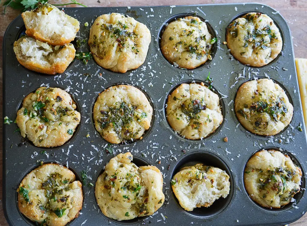 A muffin tin filled with twelve baked savory bread rolls topped with herbs and shredded cheese; two rolls are torn open, showing their soft interior.