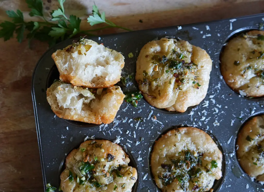 A muffin tin with baked bread rolls topped with herbs and cheese. One roll is broken open to show its soft, airy interior. Fresh parsley is in the background.