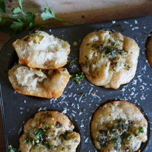 A muffin tin with baked bread rolls topped with herbs and cheese. One roll is broken open to show its soft, airy interior. Fresh parsley is in the background.