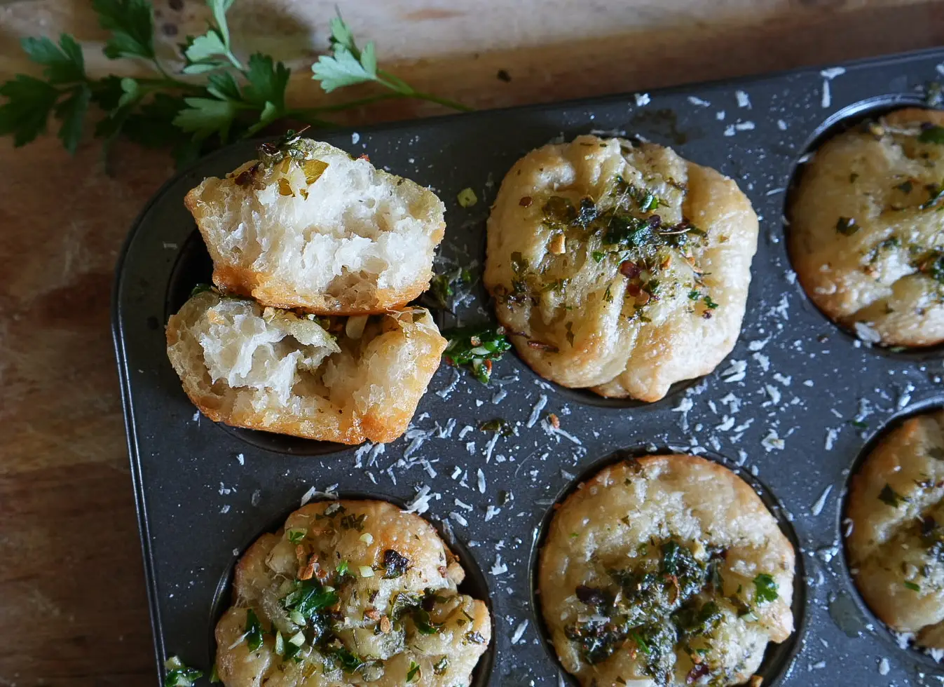 A muffin tin with baked bread rolls topped with herbs and cheese. One roll is broken open to show its soft, airy interior. Fresh parsley is in the background.