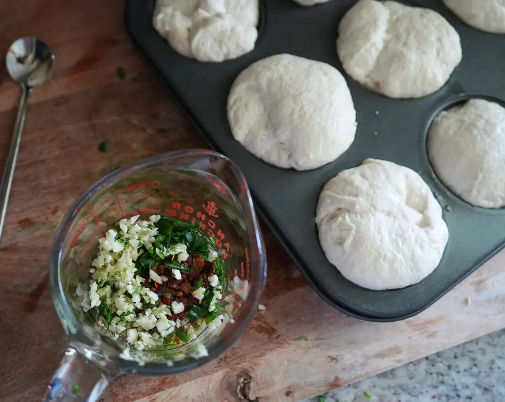 Muffin tin with dough portions next to a measuring cup containing chopped herbs, garlic, and sun-dried tomatoes on a wooden surface.