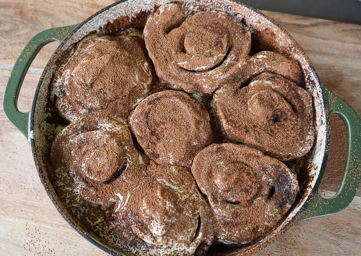 A round baking dish filled with seven large, baked cinnamon tiramisu rolls topped with a dusting of cocoa powder.