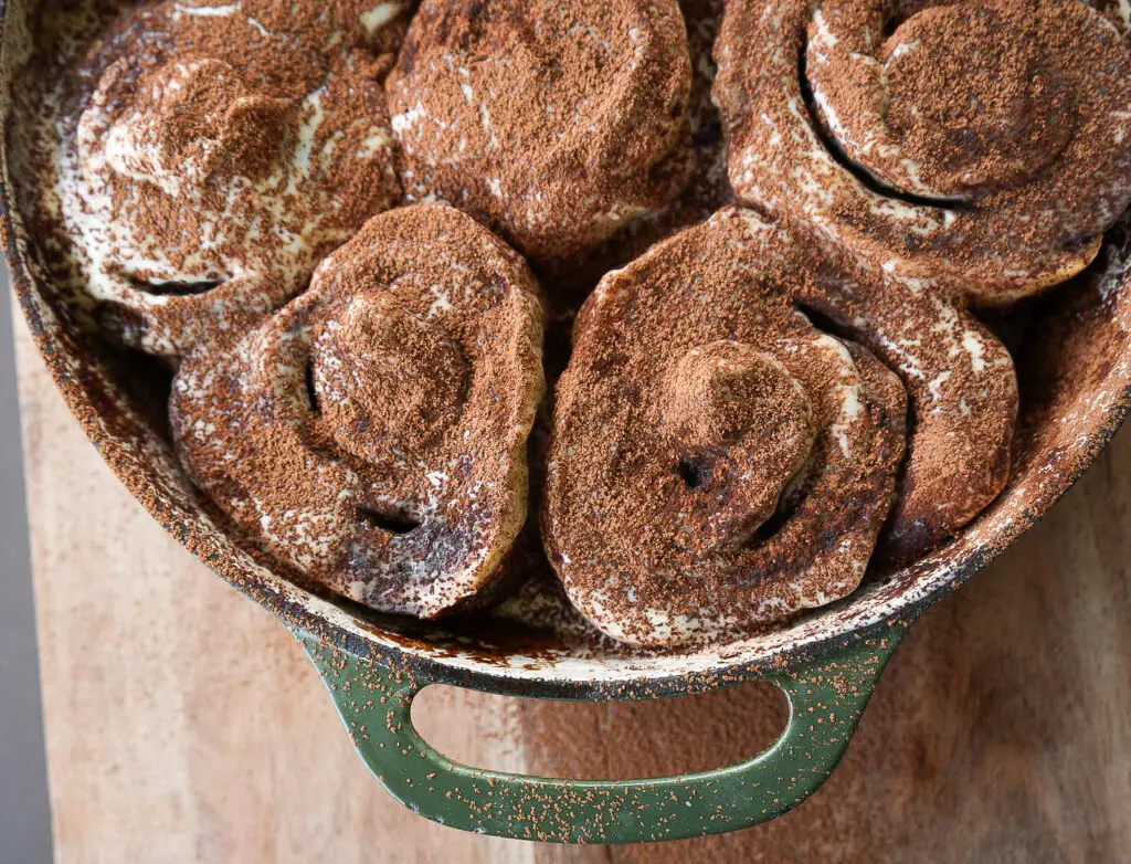 A close-up of freshly baked sourdough tiramisu rolls in a round, green baking dish, dusted with cocoa powder on a wooden surface.