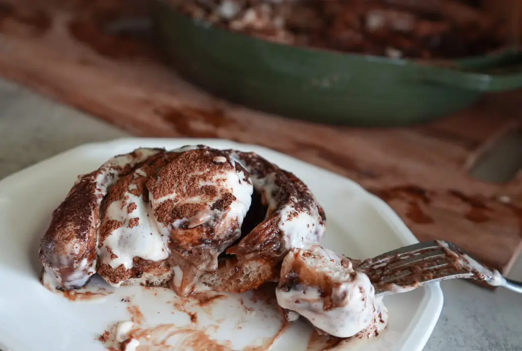 A sourdough tiramisu roll with cream cheese icing and cocoa powder sits on a plate, with a fork cutting into it; a baking dish is in the background.