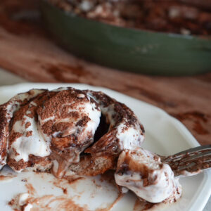 A sourdough tiramisu roll with cream cheese icing and cocoa powder sits on a plate, with a fork cutting into it; a baking dish is in the background.
