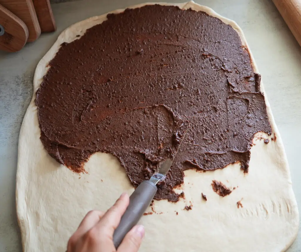 A hand spreads a layer of chocolate filling over rolled-out dough using a spatula on a light surface.