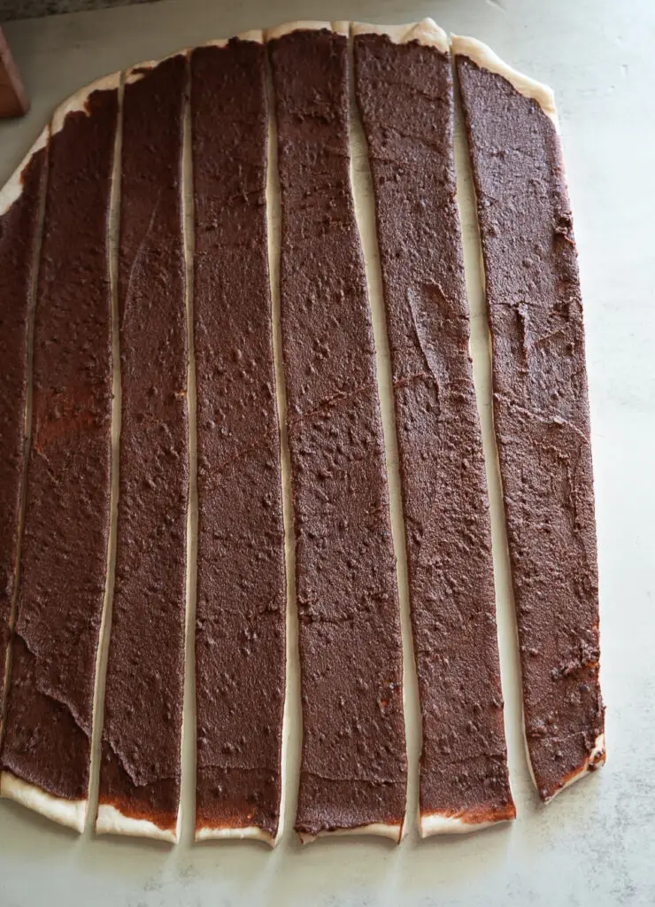 Rectangular sheet of dough spread with a brown filling and cut into long, even strips on a light surface.