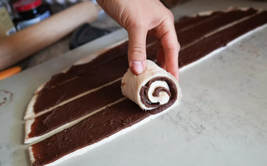 A hand rolls up a strip of dough layered with chocolate filling on a countertop, with other strips and a rolling pin visible in the background.