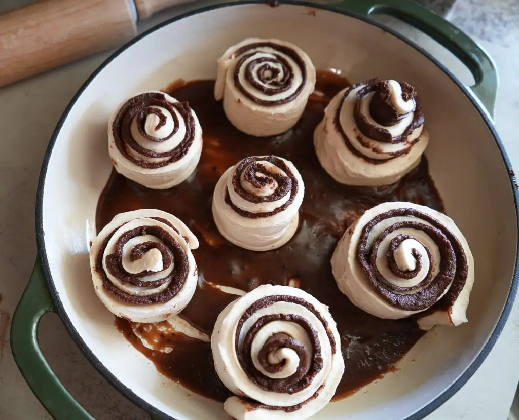 Unbaked cinnamon rolls with chocolate filling arranged in a round, green-handled baking dish, with sauce pooled at the bottom.
