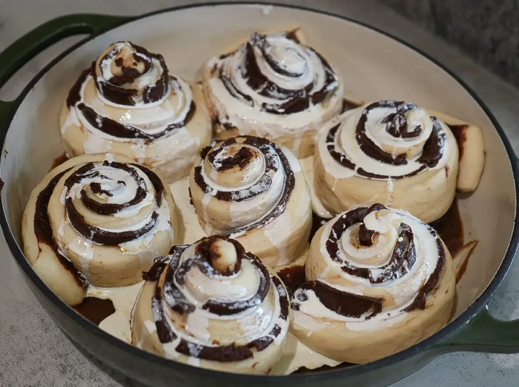 A round baking dish with seven unbaked sourdough tiramisu rolls, topped with cream or icing, sitting on a countertop.