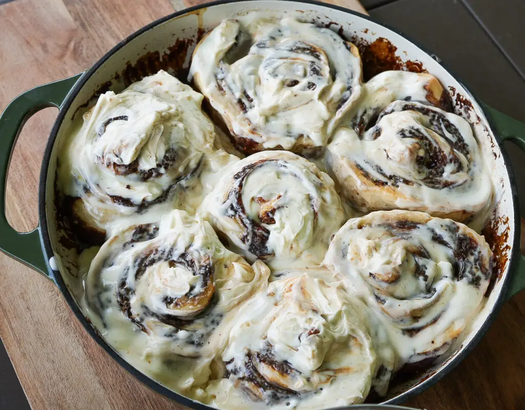 A round baking dish filled with six large sourdough tiramisu rolls topped with melted cream cheese icing, sitting on a wooden surface.