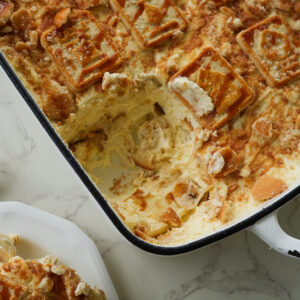 A baking dish of banana pudding with cookies on top, partially served, sits on a marble surface next to a plate with a portion and a spoon.