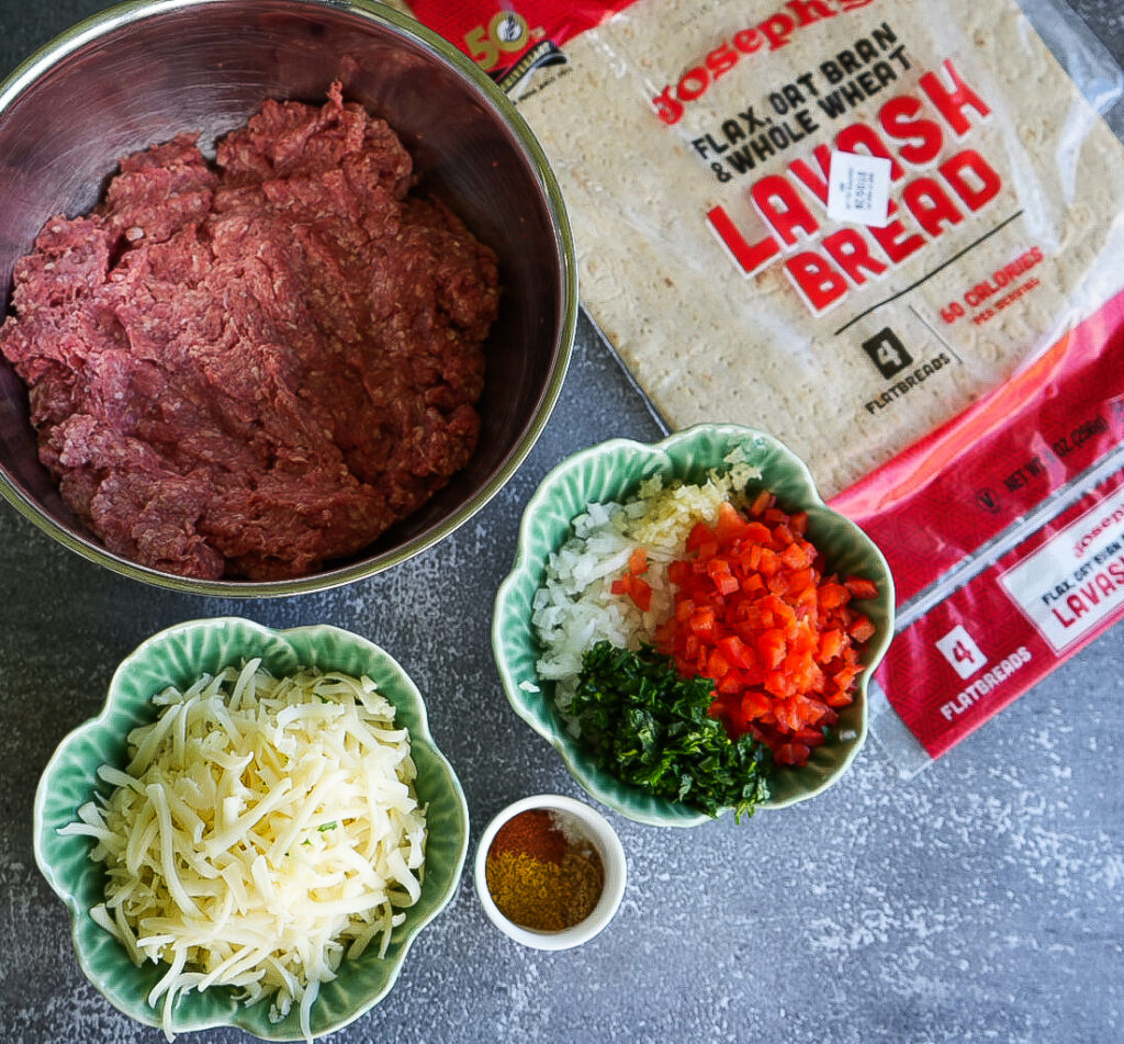 Ground beef, Lavash bread, shredded cheese, chopped onions, red bell pepper, parsley, and a small bowl of spices arranged on a gray surface.