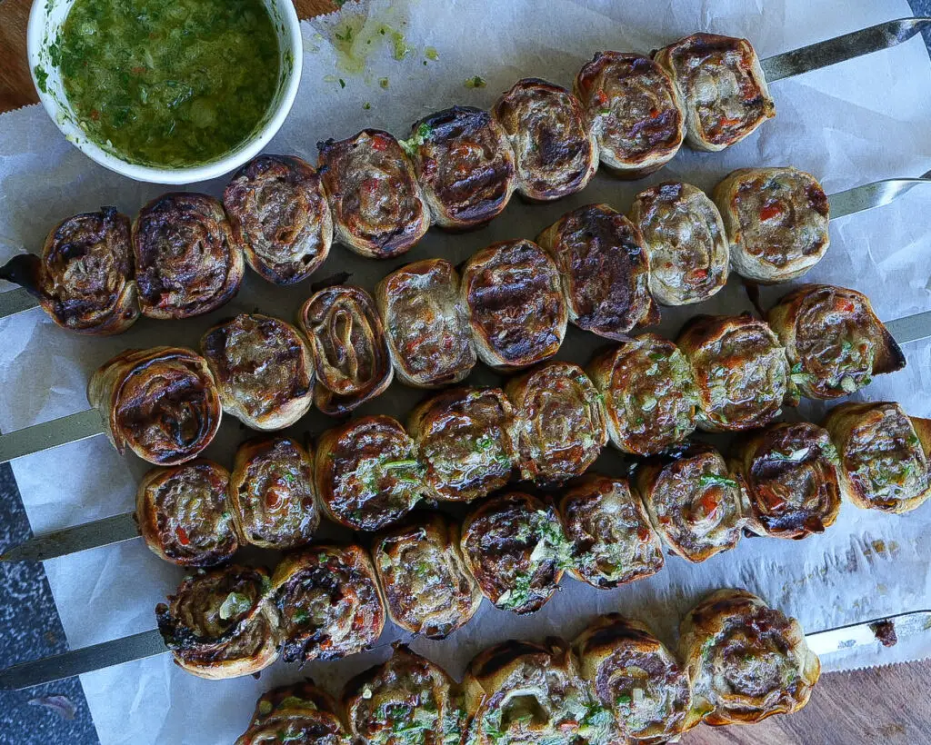 Five skewers of grilled spiral pastry rolls with visible herbs and vegetables are arranged on parchment paper next to a small bowl of green dipping sauce.
