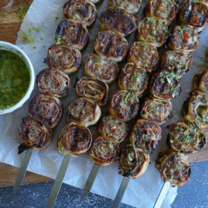Four metal skewers hold grilled spiral pastries with herbs, served on parchment paper next to a bowl of green dipping sauce and a spoon.