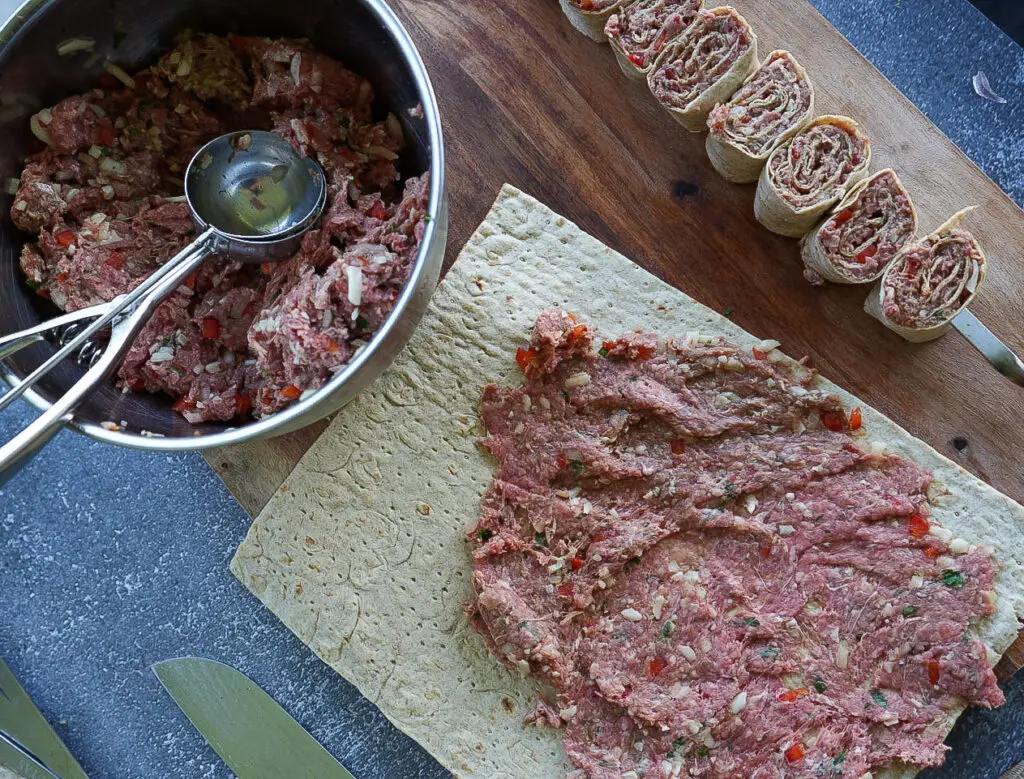 A bowl of ground meat mixture next to a flatbread being spread with the mixture, with rolled and sliced pinwheels on a cutting board.