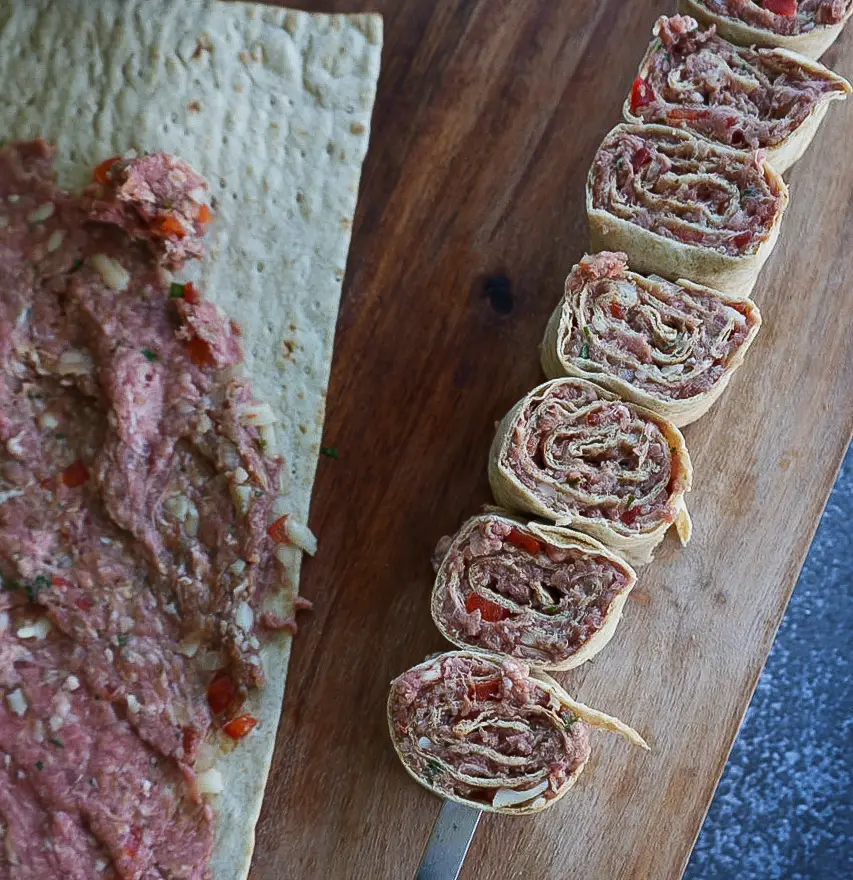 Flatbread with a spread of minced meat and vegetables is partly rolled and sliced into rounds, displayed on a wooden cutting board.