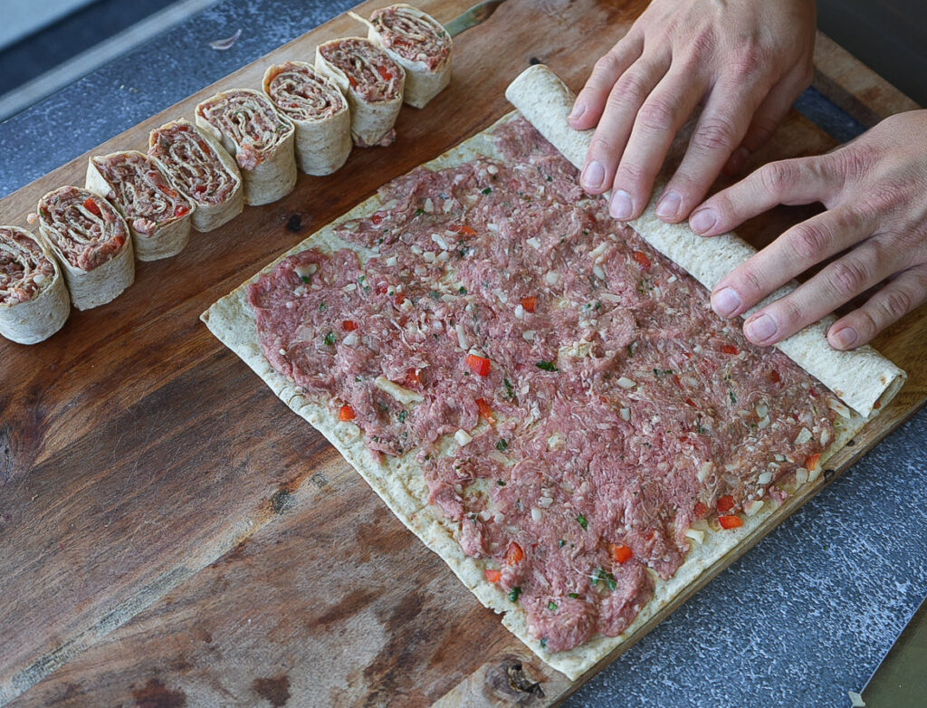 Hands rolling a flatbread topped with raw ground meat mixture; several sliced pinwheel pieces are arranged nearby on a wooden board.