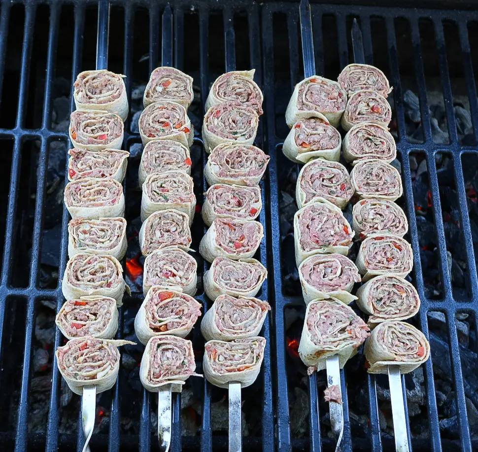 Four metal skewers with rolled flatbread and meat slices are grilling on a barbecue, with glowing charcoal visible underneath the grill grates.