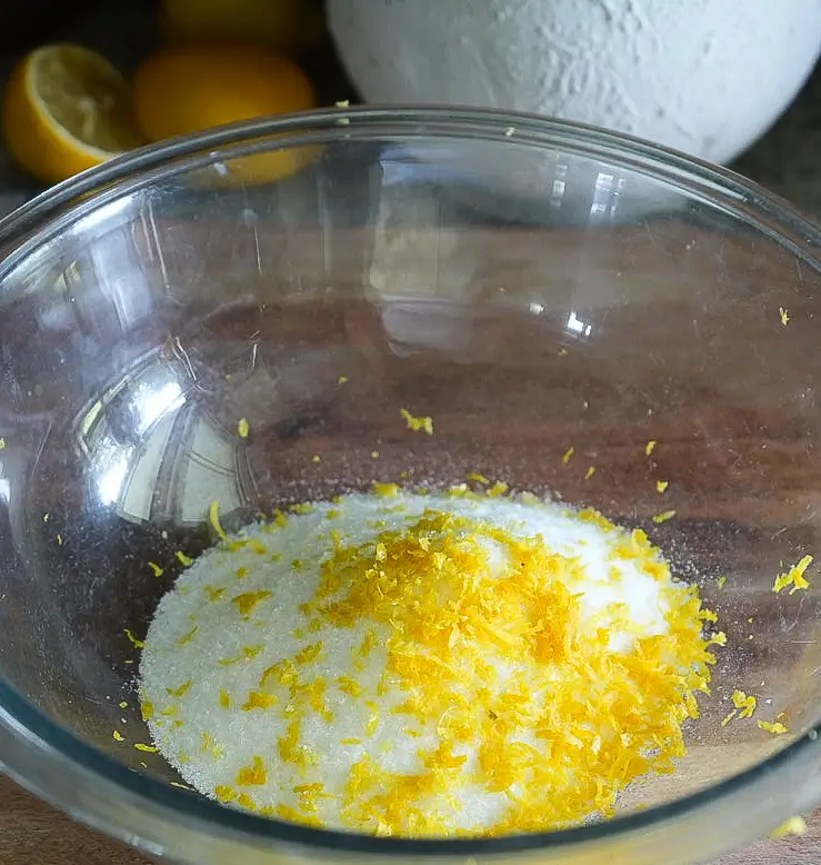 A glass bowl containing granulated sugar mixed with fresh lemon zest, placed on a wooden surface with lemons in the background.