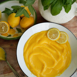 A bowl of lemon curd garnished with lemon slices sits on a wooden surface next to whole and sliced lemons and a potted plant.