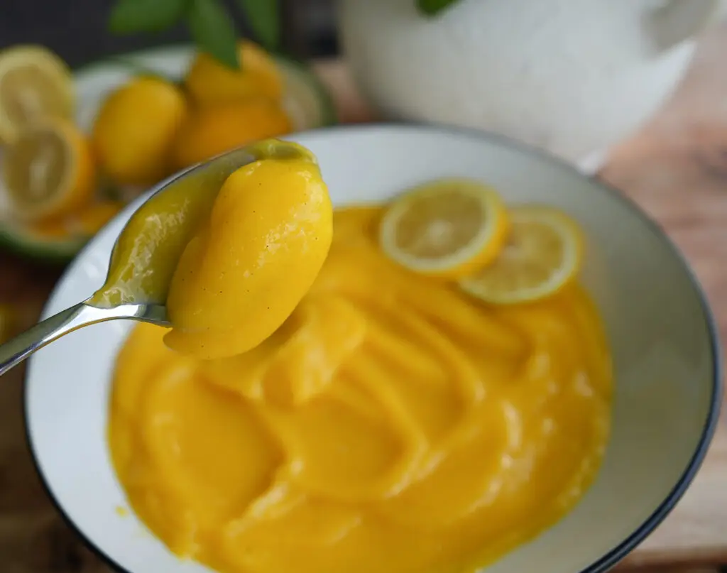 A spoon holds a portion of thick yellow mango puree above a bowl filled with the same puree, garnished with lemon slices.