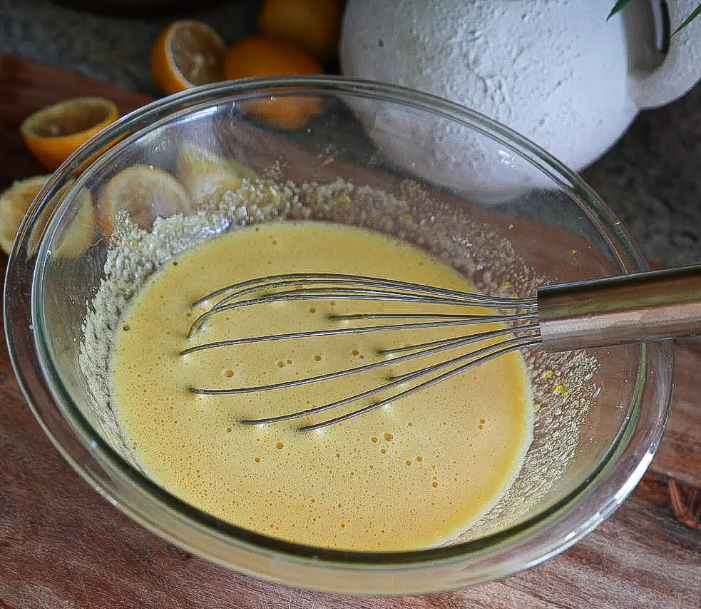 A metal whisk rests in a glass bowl of yellow batter on a wooden surface, with lemons and a white ceramic pot in the background.