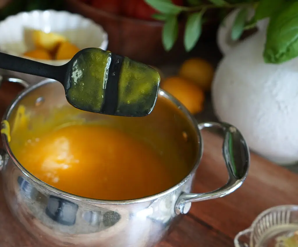 A spatula with yellow sauce rests above a metal pot containing more of the sauce, set on a wooden surface with fruit and greenery in the background.