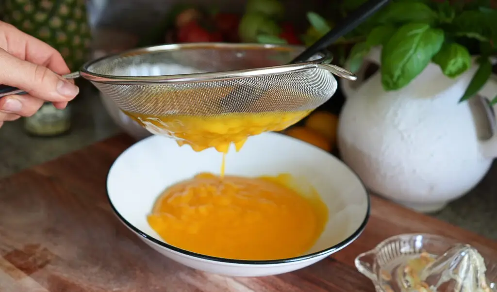 A hand holds a fine mesh strainer over a bowl, sieving yellow liquid, with kitchen items such as a white ceramic pot and fruit visible in the background.