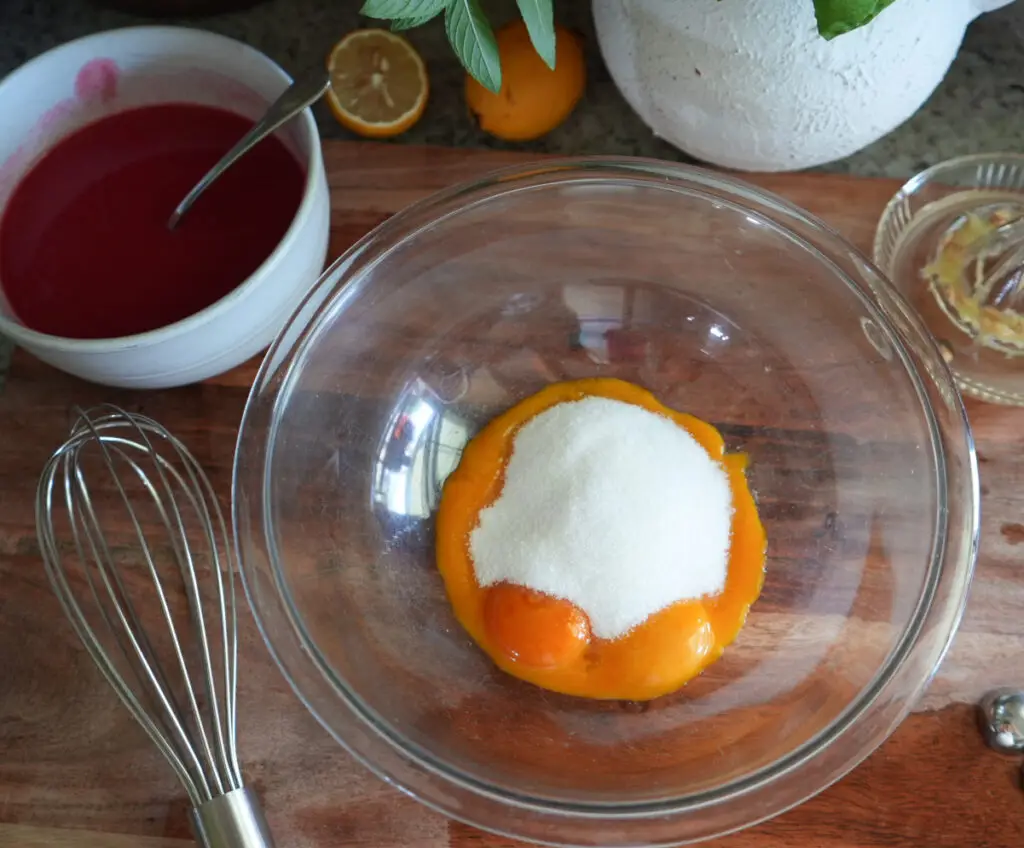 A glass bowl with egg yolks and sugar sits on a wooden surface beside a whisk, a bowl of red liquid, a squeezed lemon half, and some potted herbs.