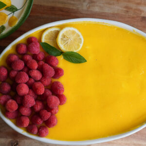 Oval dish with yellow dessert topped on one side with fresh raspberries, lemon slices, and a mint sprig; wooden surface underneath.