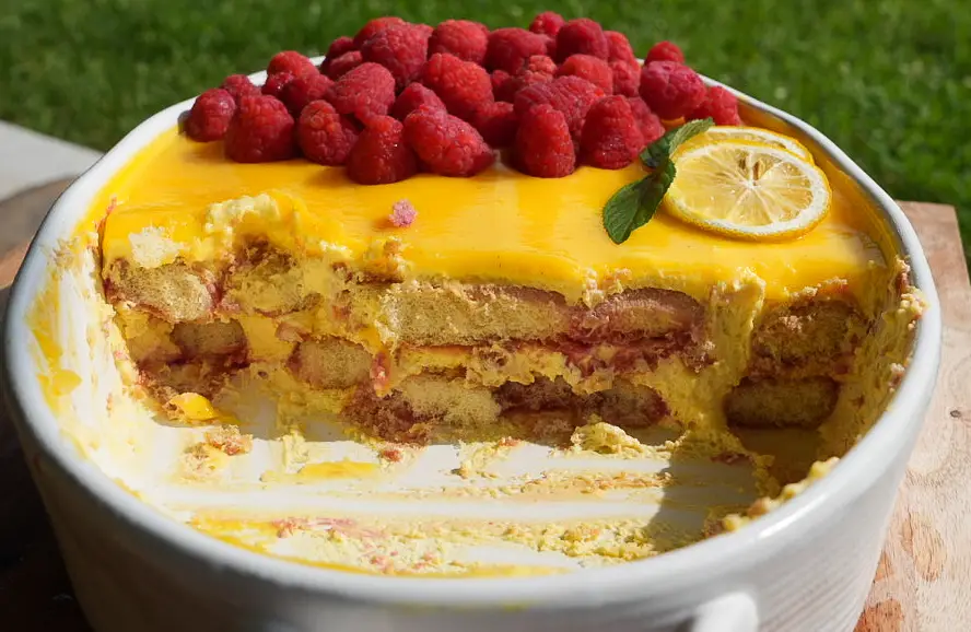 A layered dessert topped with raspberries, a lemon slice, and mint in a white dish, with a portion removed to show its creamy interior.