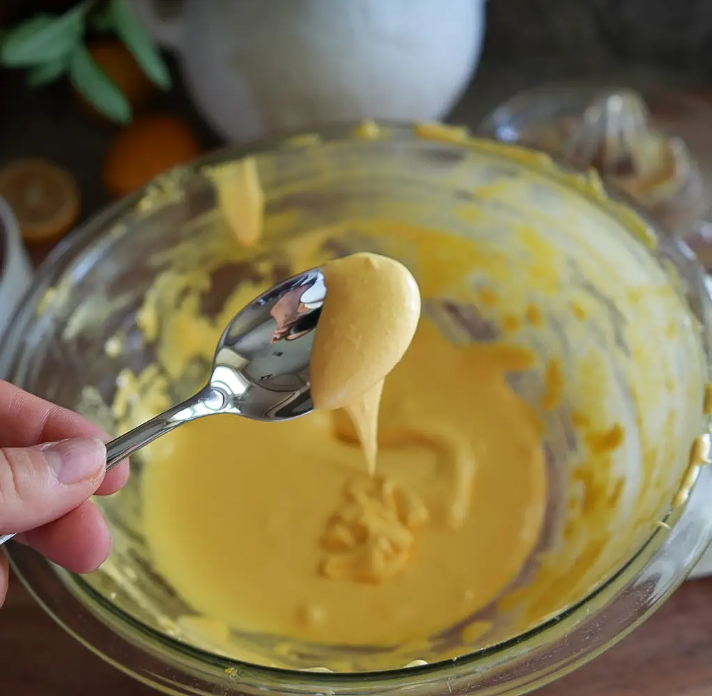 A hand holds a spoon with yellow creamy batter over a glass mixing bowl containing more of the same mixture.
