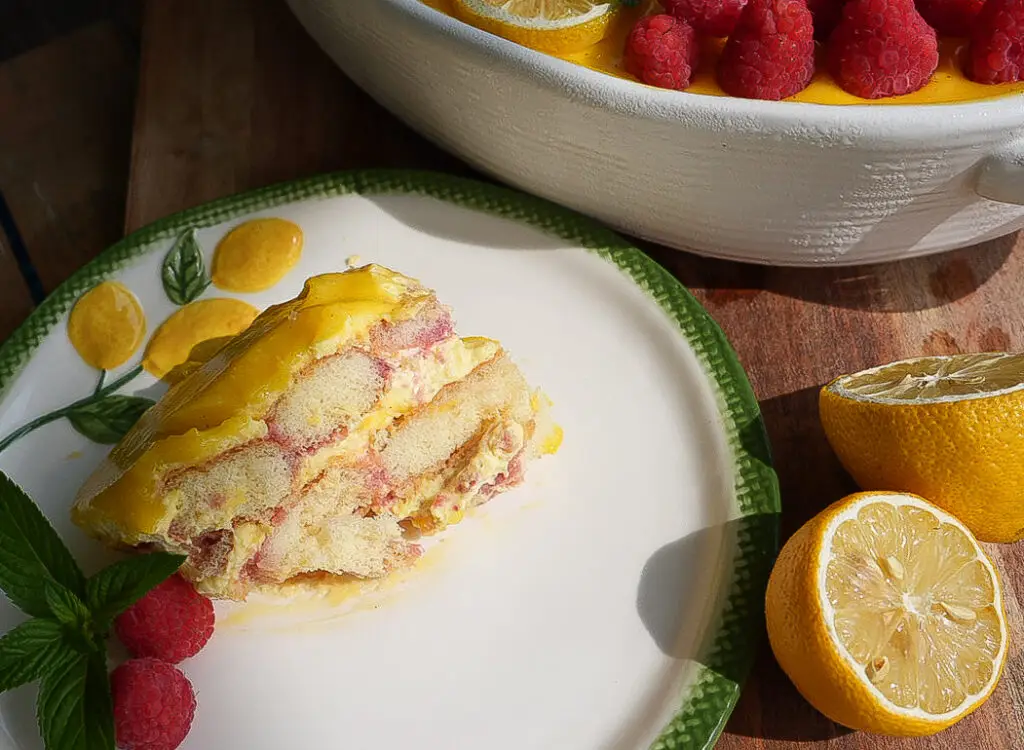A slice of raspberry and lemon dessert on a plate with fresh raspberries, mint, and lemon halves nearby; the rest of the dessert is in a dish in the background.